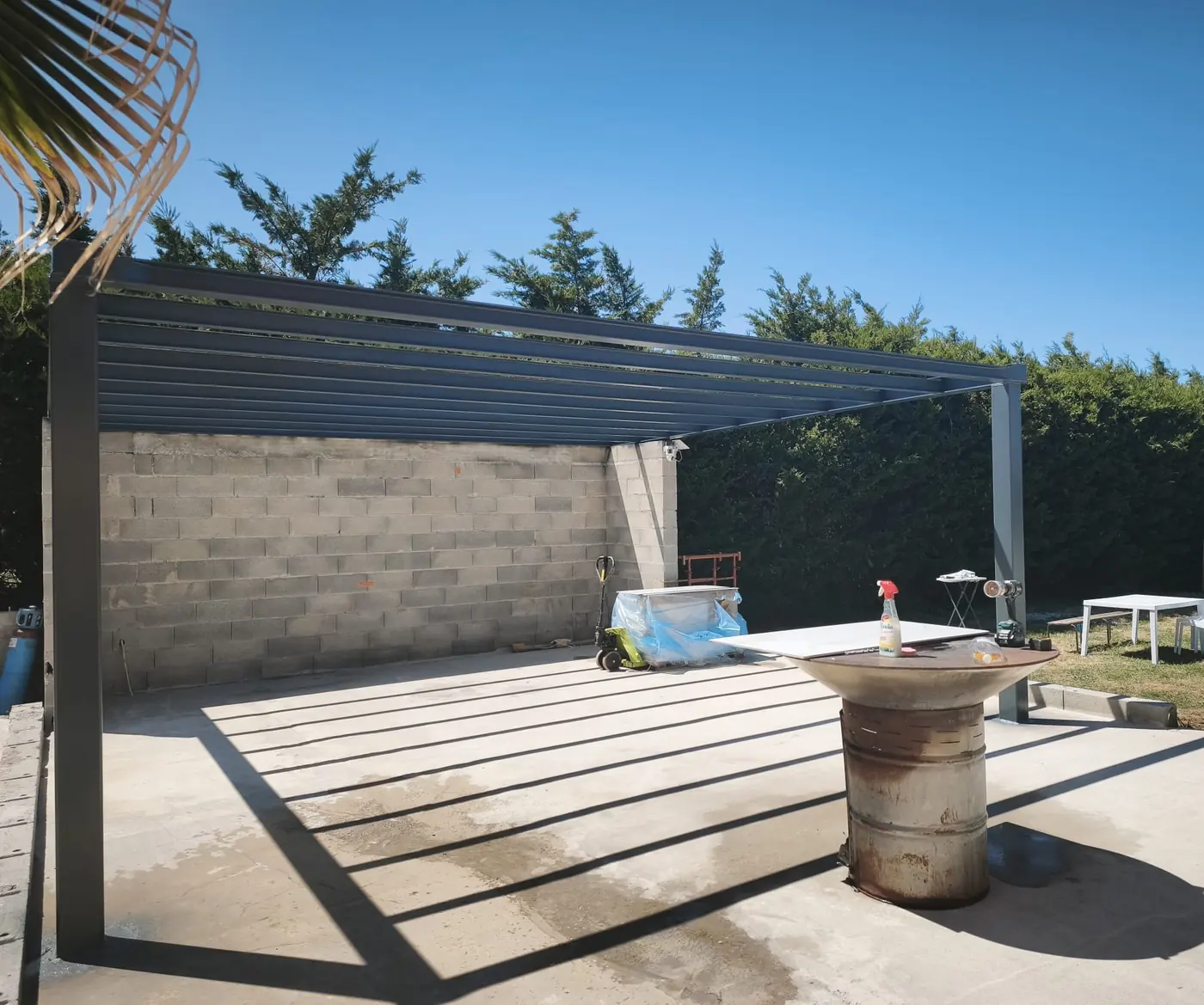 Pergola en aluminium à lames posée sur la dalle béton, murs en parpaing du pool house à l’arrière